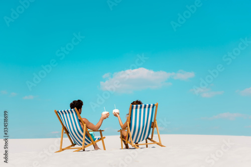 Leinwand Poster back view of young couple sitting in deck chairs with coconut drinks on sandy be