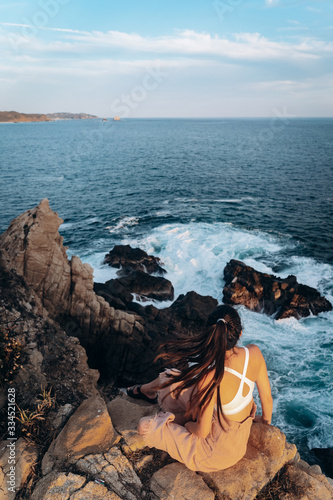 Fotografi Girl sitting on a cliff of the Pacific Ocean at sunset
