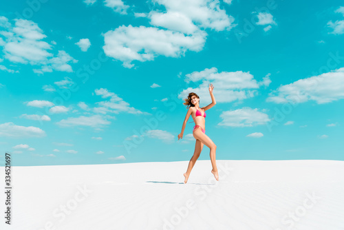 smiling beautiful sexy girl in swimsuit jumping on sandy beach with blue sky and clouds