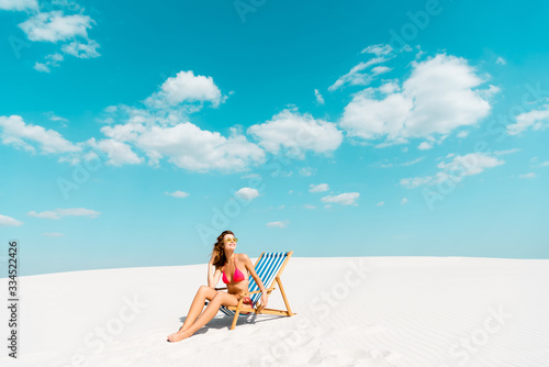 smiling beautiful sexy girl in swimsuit and sunglasses sitting in deck chair on sandy beach with blue sky and clouds