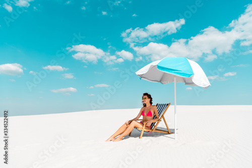 smiling beautiful sexy girl in swimsuit and sunglasses sitting in deck chair under umbrella on sandy beach