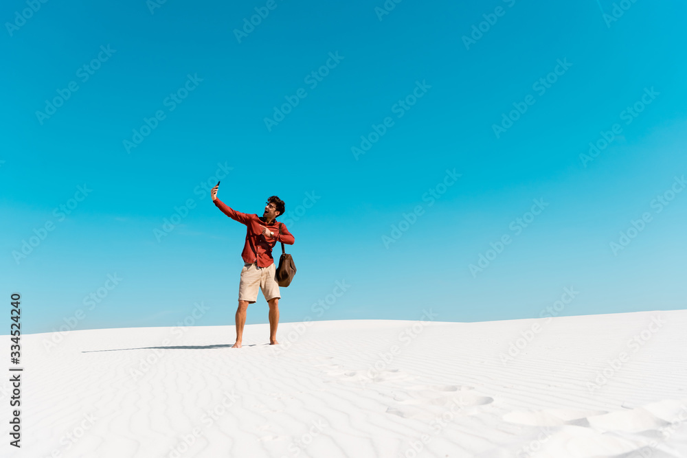handsome man with leather bag taking selfie on smartphone on sandy beach against clear blue sky