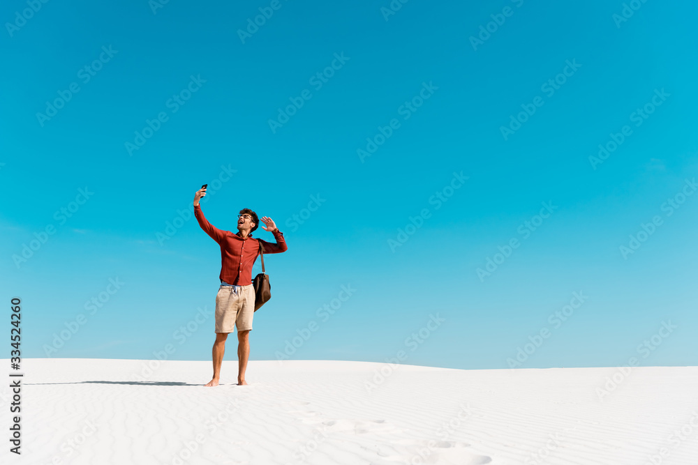 handsome man with leather bag using smartphone on sandy beach against clear blue sky