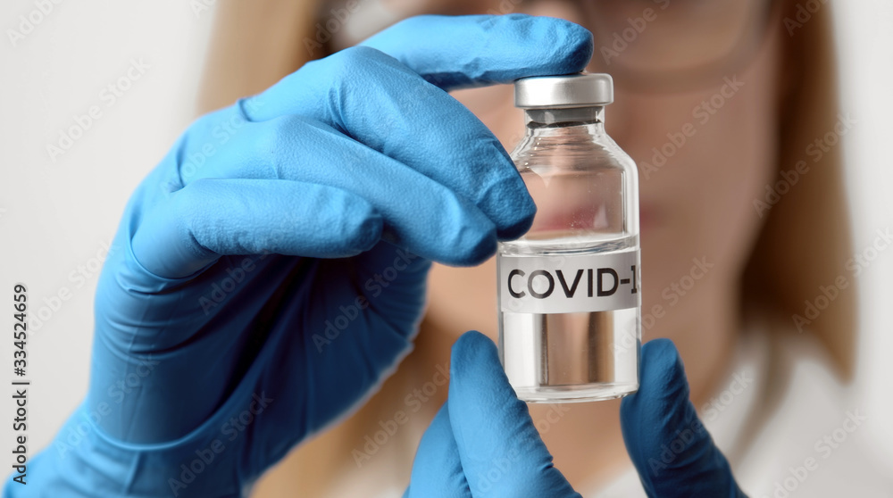 Close-up of doctor's hands in gloves holding vaccine bottle with COVID-19 inscription, focus on vial Close-up of doctor's hands in gloves holding vaccine bottle with COVID-19 inscription, focus on vial