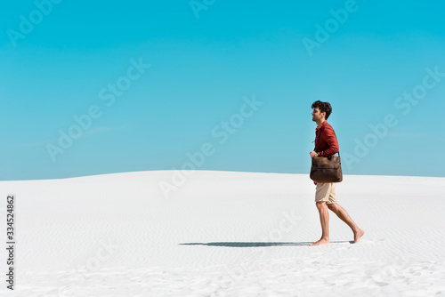 side view of handsome man with leather bag walking on sandy beach against clear blue sky