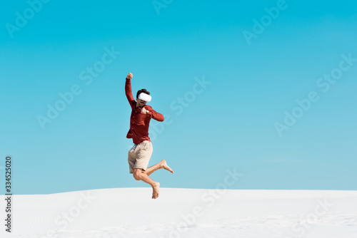 man on sandy beach in vr headset jumping against clear blue sky