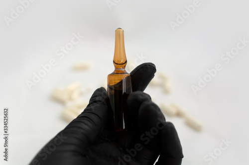 Hand in black latex glove holding medical glass ampule with medicine. Selective focus. Amber glass ampoule is held by hand in black medical glove with scattered pills at the background. Virus cure.