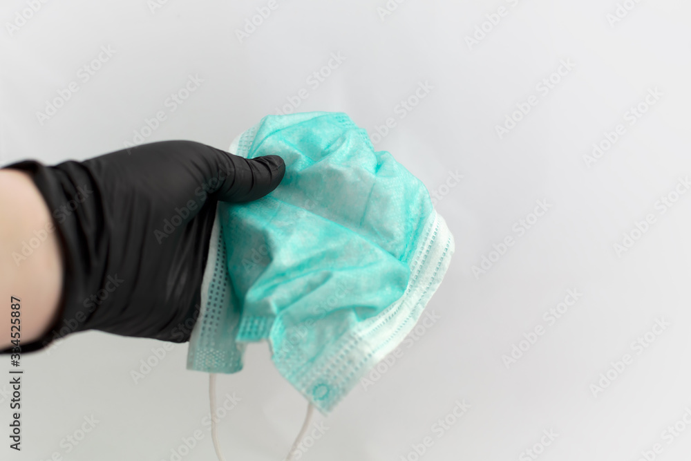 Fototapeta premium Hand in black latex glove holding green disposable medical face mask. Selective focus. Face protection mask is held by hand in black medical glove at white background. Virus care.