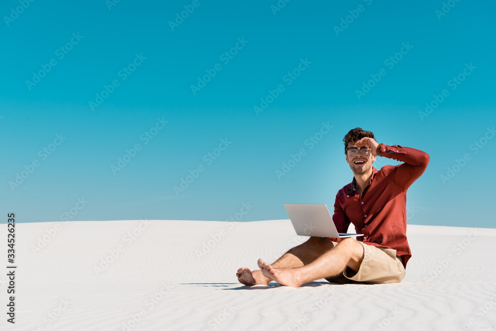 happy freelancer on sandy beach using laptop against clear blue sky