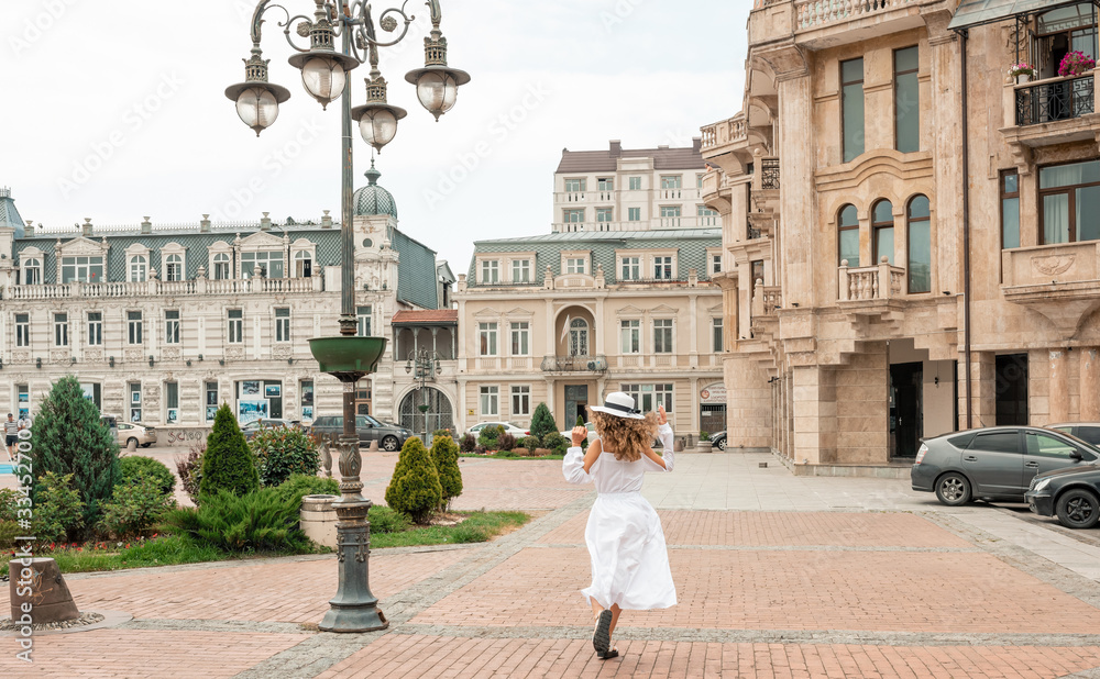 Cute girl female explore the city in Europe. Curly woman in white chiffon dress and white hat walking among famous buildings. Travel and tourism. Lifestyle. Fashion shot. Back view