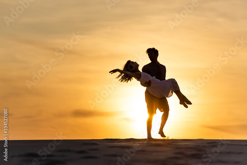 silhouettes of man spinning around woman on beach against sun during sunset