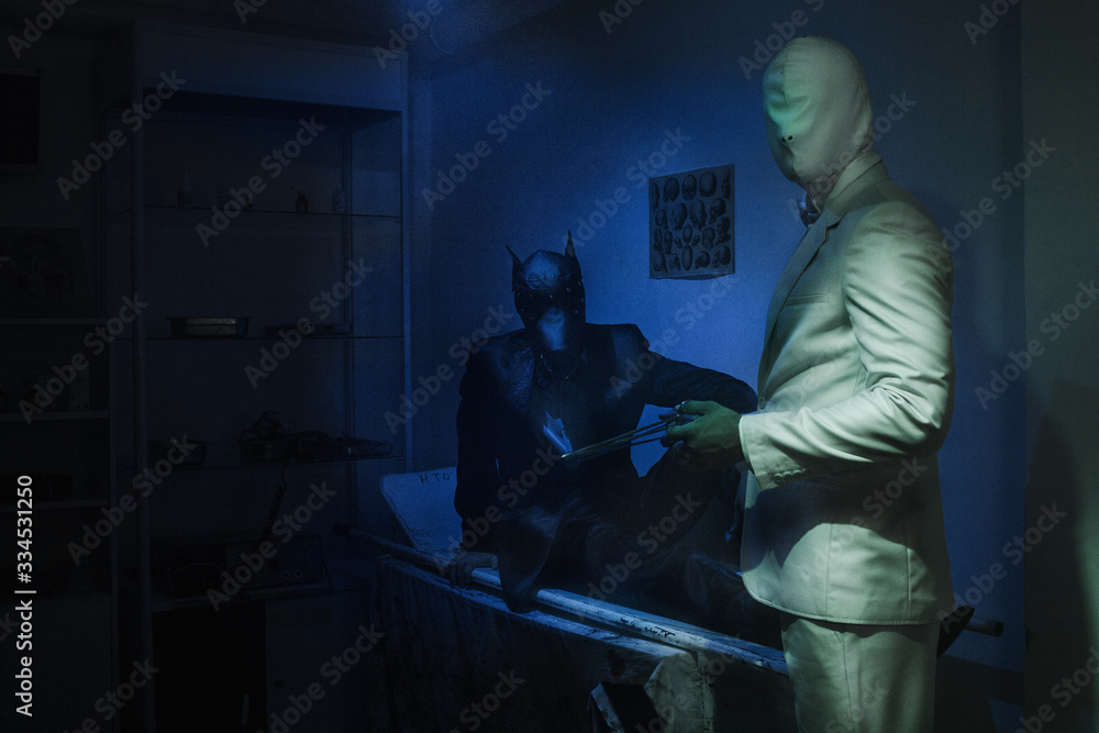 two guys, one in a black suit and a dog mask, one in a white suit with a white latex mask, is sitting on a stretcher two guys, one in a black suit and a dog mask, one in a white suit with a white latex mask, is sitting on a stretcher