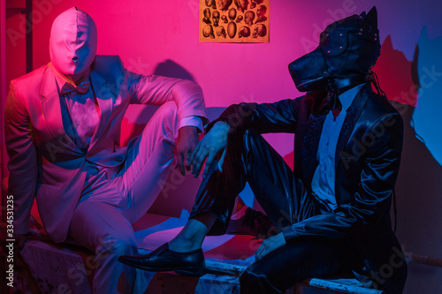 two guys, one in a black suit and a dog mask, one in a white suit with a white latex mask, is sitting on a stretcher photo