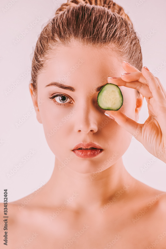 Young beautiful woman posing with cucumber slice on face isolated on background. Attractive girl beauty model with perfect clean face skin holding cucumber slice on eye. Beauty treatment, cosmetology Young beautiful woman posing with cucumber slice on face isolated on background. Attractive girl beauty model with perfect clean face skin holding cucumber slice on eye. Beauty treatment, cosmetology