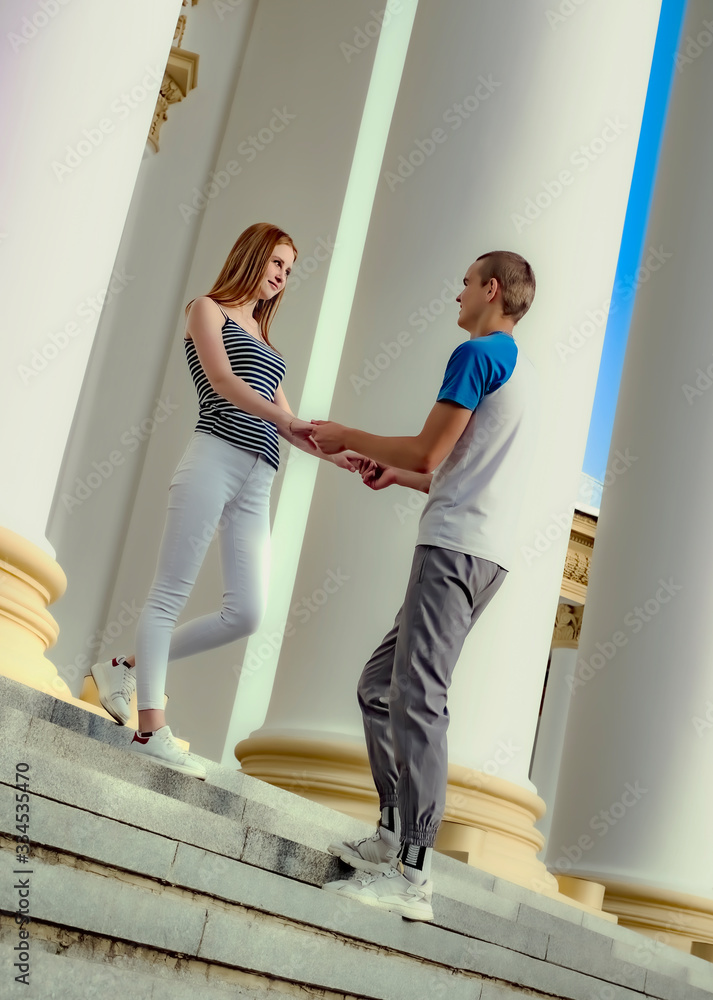 Couple hold hands and looks at each other standing on the stairs near the columns in a summer park