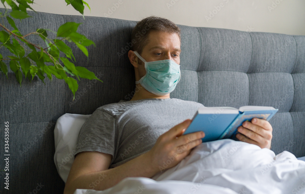 Sick man with medical mask reading book in bed. Home quarantine concept.