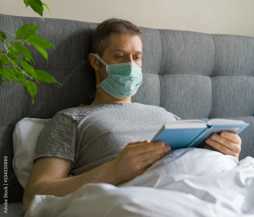 Sick man with medical mask reading book in bed. Home quarantine concept. Sick man with medical mask reading book in bed. Home quarantine concept.