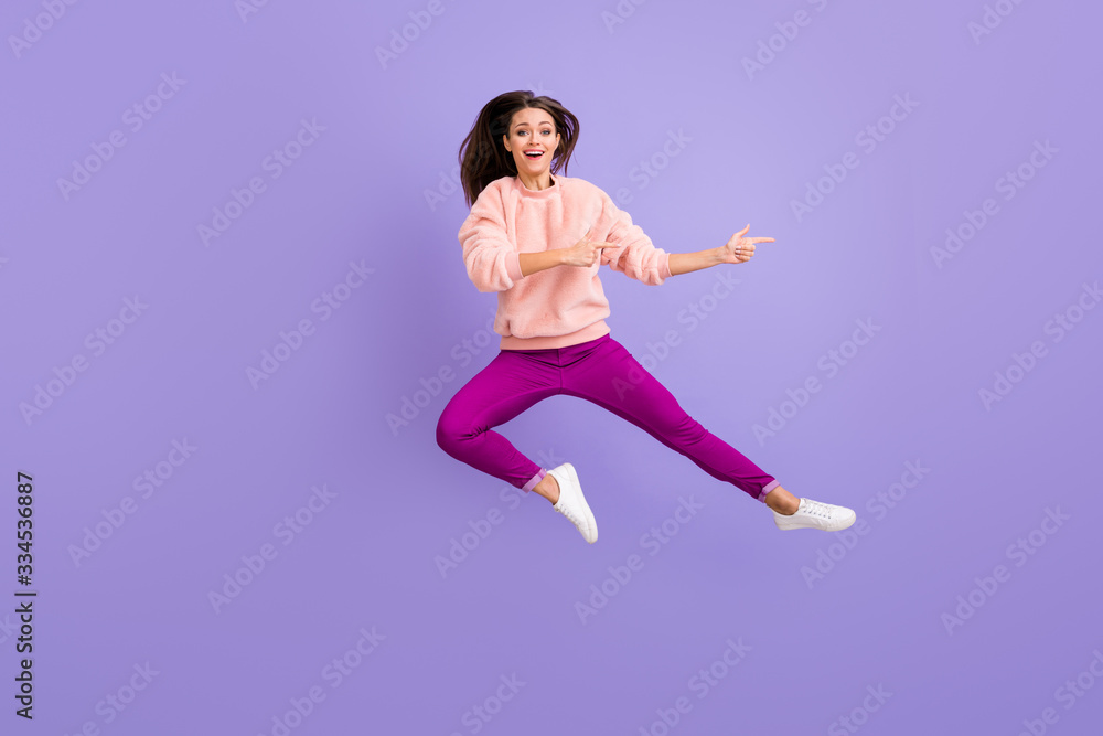 Full length photo of cheerful lady jumping high indicating fingers empty space amazing sale wear casual fluffy pullover pants shoes isolated purple color background Full length photo of cheerful lady jumping high indicating fingers empty space amazing sale wear casual fluffy pullover pants shoes isolated purple color background