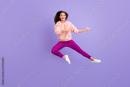 Full length photo of cheerful lady jumping high indicating fingers empty space amazing sale wear casual fluffy pullover pants shoes isolated purple color background