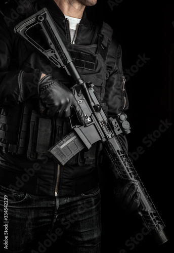 A man holds an automatic carbine with a collimator sight on a dark back. A fighter in dark clothing and a tactical vest with a weapon in his hands. Poster concept for police, security or military.