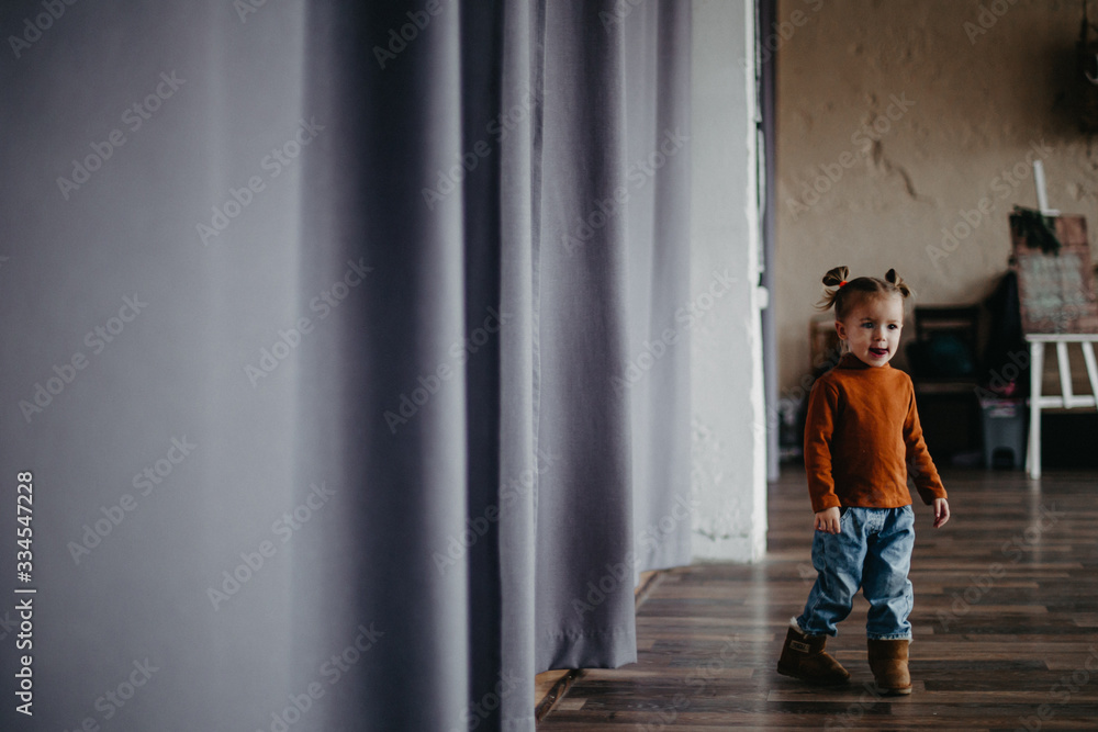 little attractive girl of 2 years is having fun and posing for a photo in a large studio with curtains by the window little attractive girl of 2 years is having fun and posing for a photo in a large studio with curtains by the window