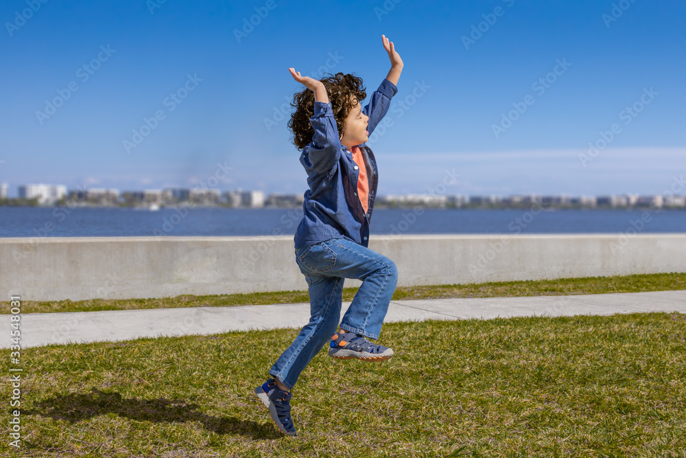 Fototapeta premium An excited little boy hops and skips in the air, arms and legs up along the waterfront.