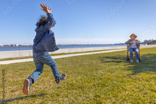 An excited toddler leaps in the air runs towards grandma sitting in a wheelchair at the park The handicap grandmother in the distance awaits for her happy grandson jumping in the air with arms up.