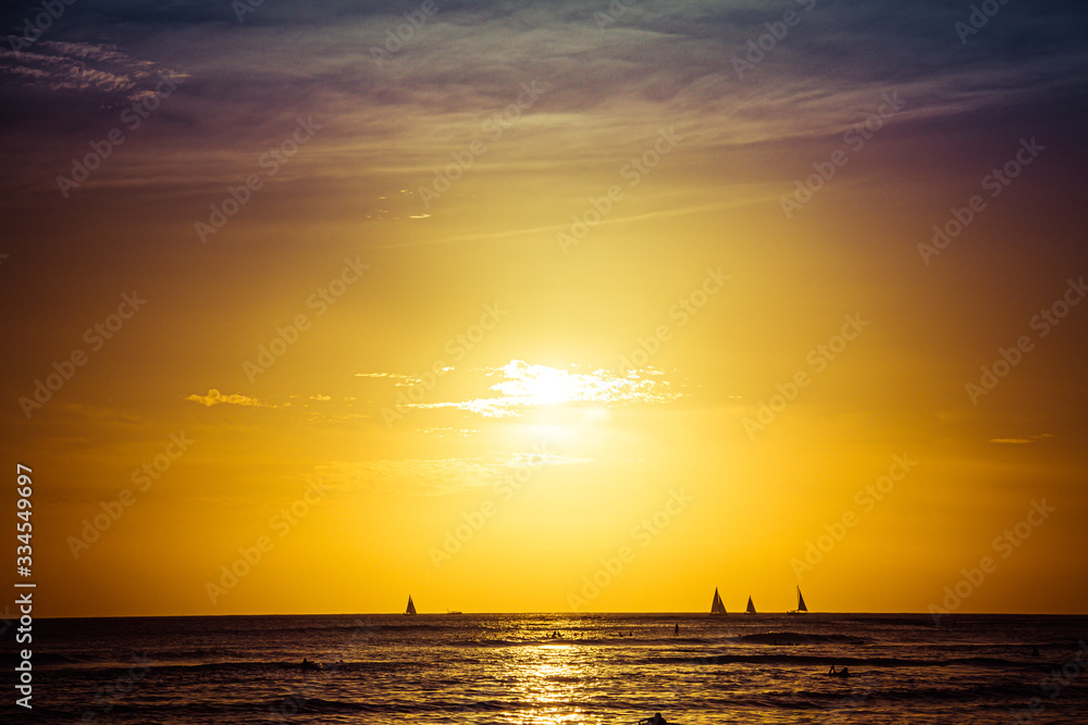 Surfers and sail boats in the ocean during sunset in the Hawaiian Islands.