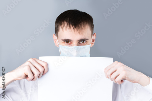 Man in a medical mask. A man in a white shirt, looks at the camera and holding a sheet of clean white paper, copy space, space for text. Chinese Coronavirus , Covid-19