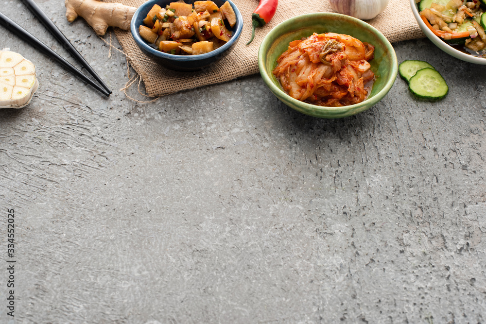 high angle view of bowls with kimchi near chopsticks, ginger, chili pepper, sliced cucumber and garlic on concrete surface high angle view of bowls with kimchi near chopsticks, ginger, chili pepper, sliced cucumber and garlic on concrete surface