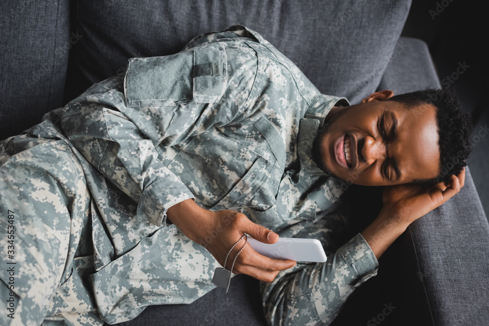 Fototapeta premium lonely african american soldier crying while holding army badge and smartphone, suffering from PTSD at home