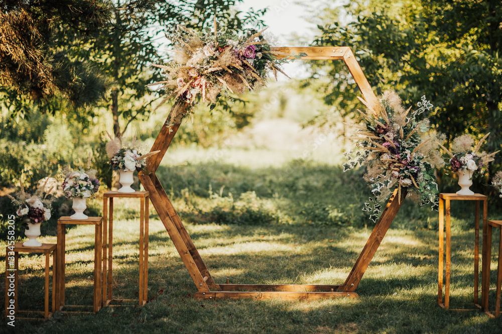 Wedding wooden arch in rustic style decorated with grass hay field color and flowers. Near wooden boxes with flower bouquets. Wooden arch and flower decoration. Wedding ceremony in sunny summer day.