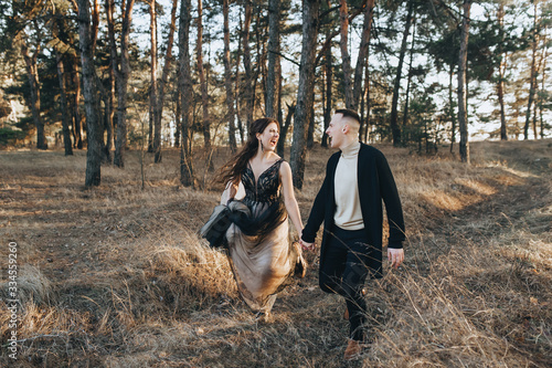Loving groom in a black cardigan and a bride in an expensive dress run, scream and hold hands in the woods in nature. Wedding portrait of cheerful and smiling newlyweds. Funny picture, concept. © shchus