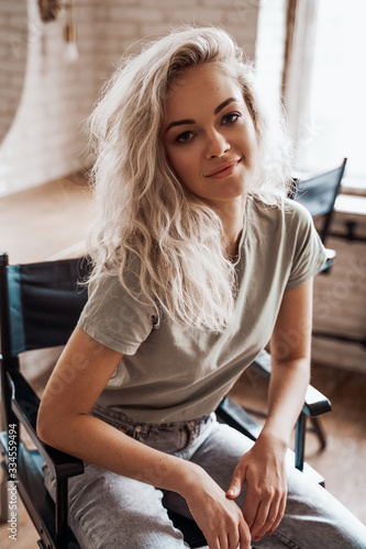 Cool and smiling young female model with blond curly hair and white boots posing on a make up chair in a bright room in front of mirrors, looking cute and interested