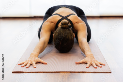Yogi woman in balasana over bright white background. Hands of lady extended on pink mat and body resting on legs at studio. Healthy lifestyle, stretching exercise, wellness, yin yoga concepts photo
