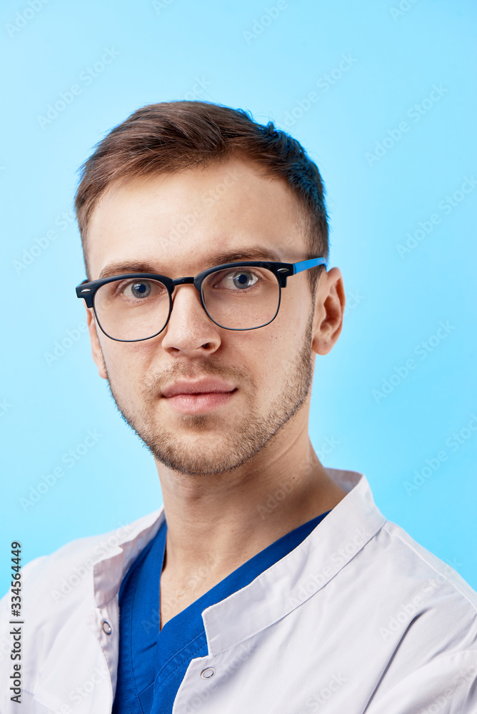 Portrait of intern doctor in white medical coat and glasses with a serious face expression isolated on blue wall background Portrait of intern doctor in white medical coat and glasses with a serious face expression isolated on blue wall background