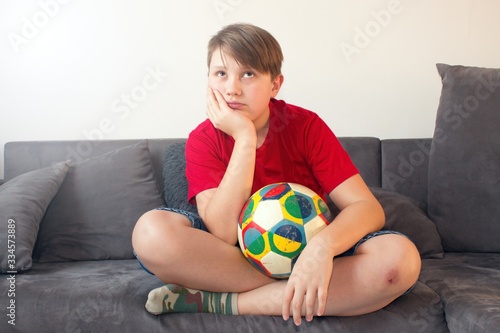 Bored boy sitting on the couch and holding a football ball.Boredom in home sandbox. While it`s not always fun, stay at home. Coronavirus pandemic. Protection against covid infection 19.
