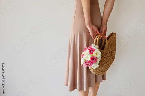 Conceptual image of a woman holding fashionable wicker bag with spring flowers. Female with colorful white and pink ranunculus bouquet over white background. Close up, copy space, cropped shot.