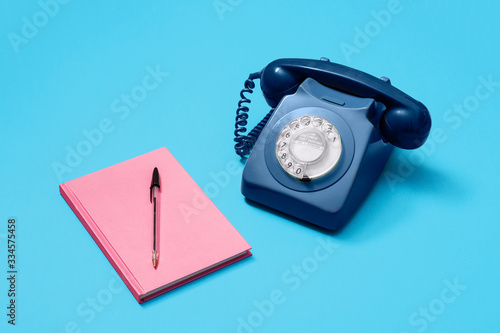 Blue vintage antique rotary phone on a blue background with a pink notebook and pen.