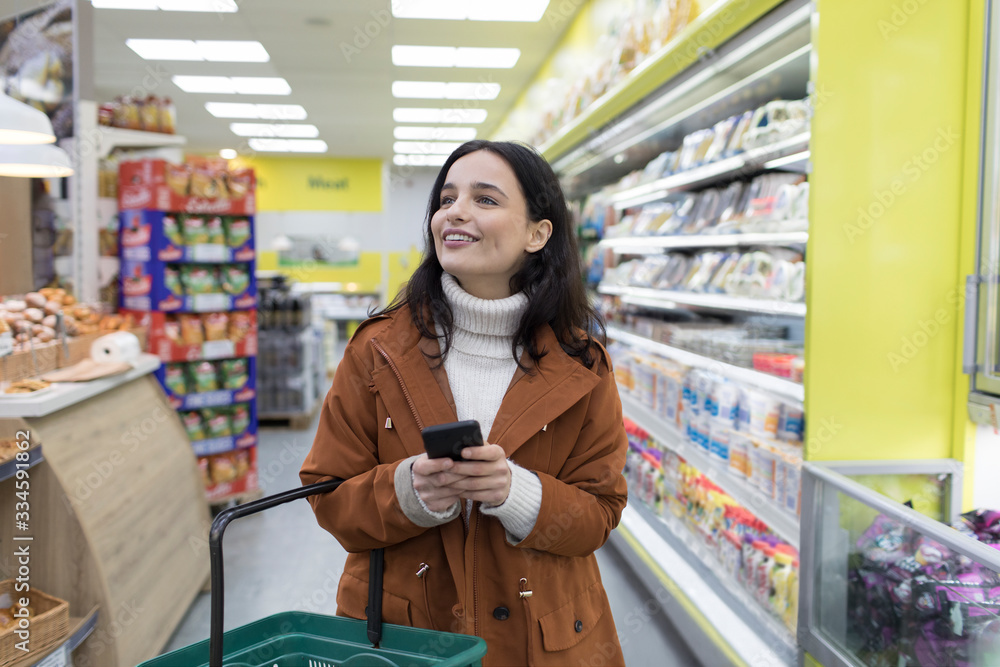 © Sam Edwards/Caia Image - Smiling young woman with smart phone grocery shopping in supermarket © Sam Edwards/Caia Image - Smiling young woman with smart phone grocery shopping in supermarket