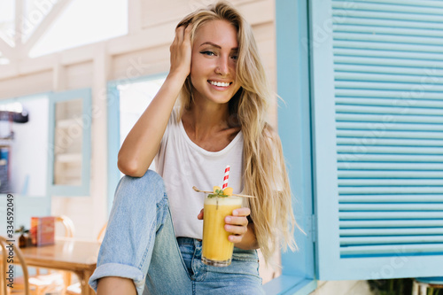 Glamorous long-haired lady drinking cocktail with pleasure. Photo of positive european woman holding glass of cold beverage.