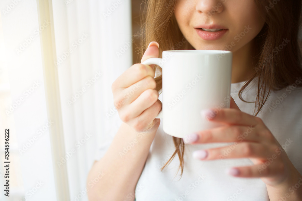 Young woman on kitchen during quarantine. Stand at window with white curtains and hold cup with tea or coffee. Drinking alone. Calm peaceful woman. Cut view. Young woman on kitchen during quarantine. Stand at window with white curtains and hold cup with tea or coffee. Drinking alone. Calm peaceful woman. Cut view.