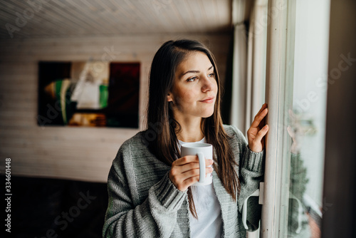 Young woman spending free time home.Self care,staying home.Enjoying view,gazing through to the window.Quarantined person indoors.Serene mornings.Avoiding social contact.