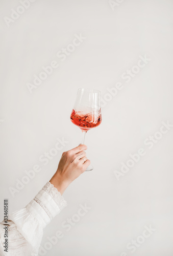 Womans hand in white shirt holding and turning glass of rose wine over white wall background. Wine shop, wine tasting, bar, wine list concept
