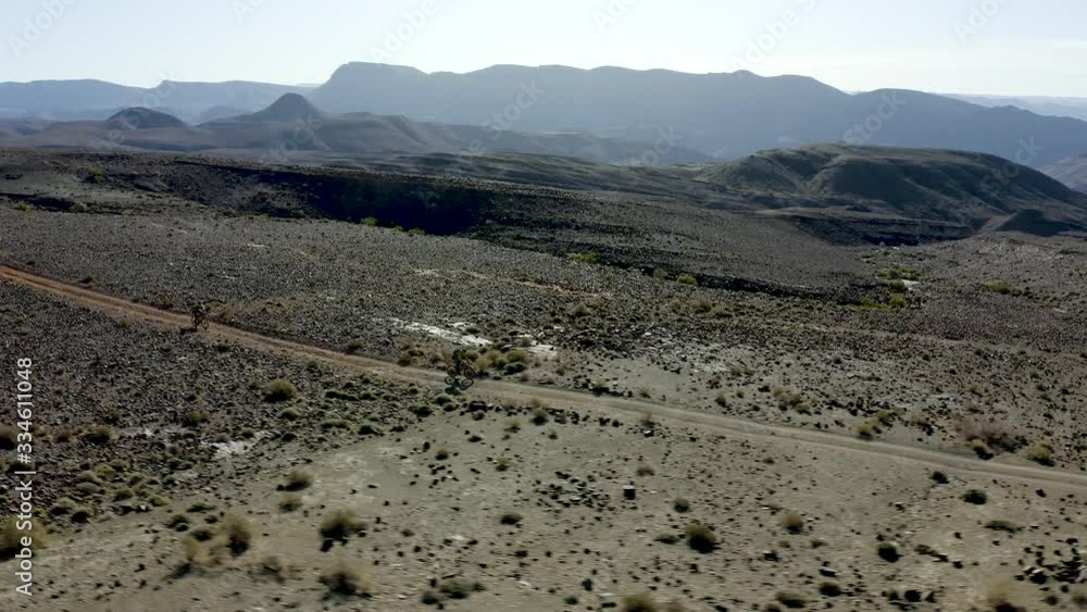 An aerial POI tracking two cyclists descending a barren desert ridge, bikepacking through a vast expanse of arid sand and rock and stark mountain ridges stretching as far as the eye can see. An aerial POI tracking two cyclists descending a barren desert ridge, bikepacking through a vast expanse of arid sand and rock and stark mountain ridges stretching as far as the eye can see.