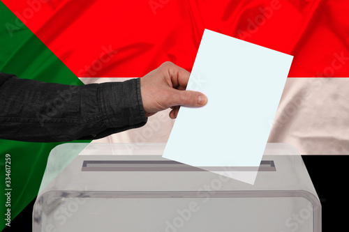 male voter drops a ballot in a transparent ballot box on the background of the national flag of Sudan, concept of state elections, referendum