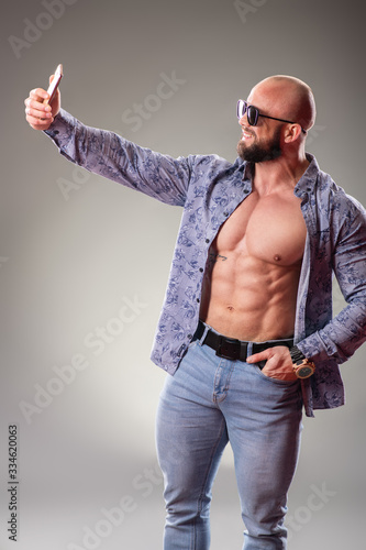 Sexy unbuttoned muscular bodybuider with sunglasses on the gray background. Selfie concept photo