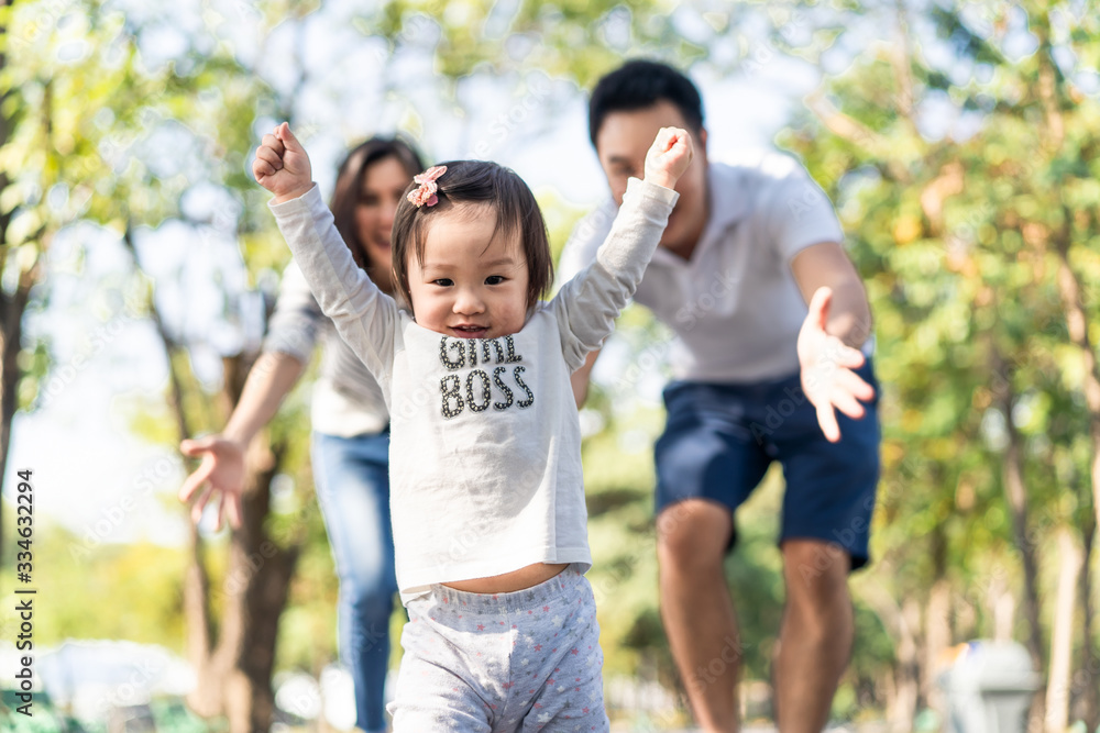 Young cute happy little Asian toddler girl running in park with parents. Mom dad taking care daughter by following looking after carefully. Kid smiling enjoy learning to walk and run, family relation.