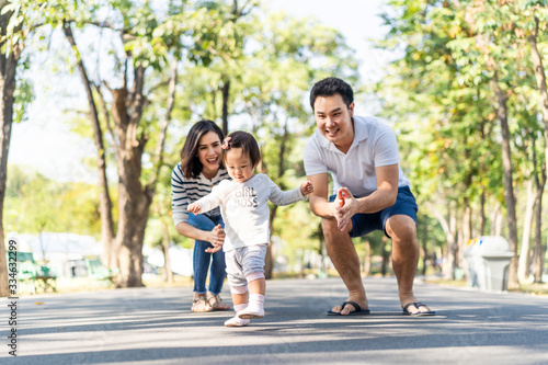 Fototapeta Naklejka Na Ścianę i Meble -  Young cute happy little Asian toddler girl running in park with parents. Mom dad taking care daughter by following looking after carefully. Kid smiling enjoy learning to walk and run, family relation.