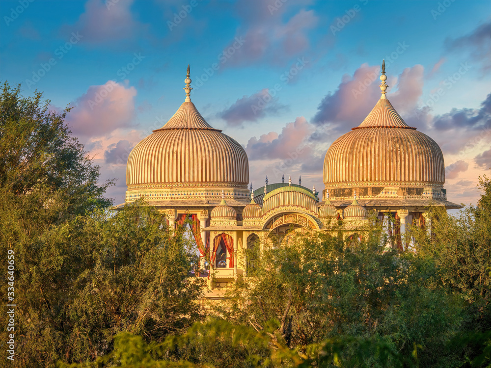 The beautiful, ornate domes and arches of a traditional North Indian palace in Mandawa, Rajasthan, which has been restored and turned into a hotel, viewed through trees and set against a sunrise sky.